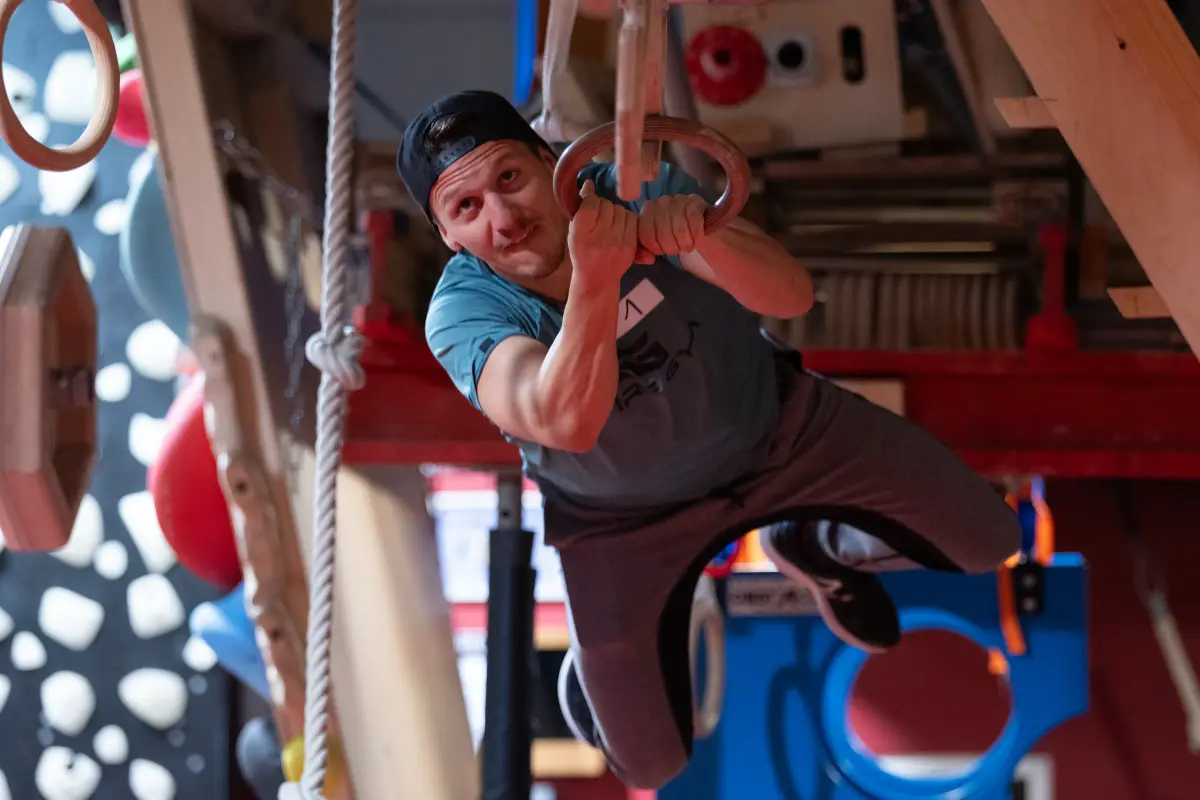 Ninja-Warrior-Athlet mit Startnummer 1 hängt an Turnringen über dem Parcours